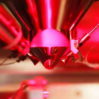 A close-up of a high-tech metallic device with a conical tip, illuminated by red lighting. The image suggests advanced scientific or quantum research equipment.