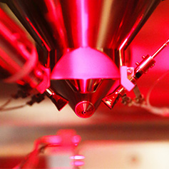 A close-up of a high-tech metallic device with a conical tip, illuminated by red lighting. The image suggests advanced scientific or quantum research equipment.